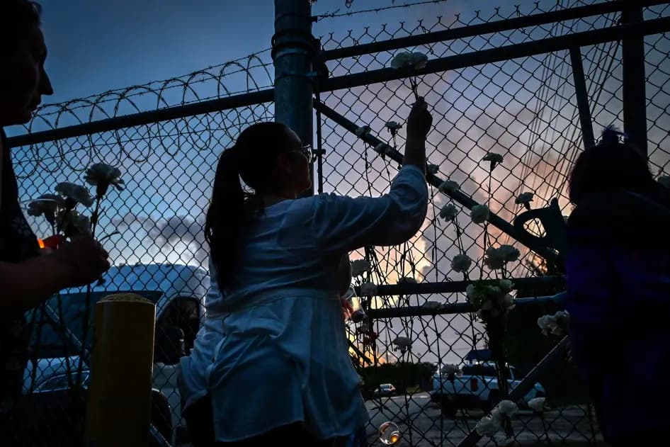 Silhouettes of vigil attendees placing white flowers into a barbed-wire fence at dusk.