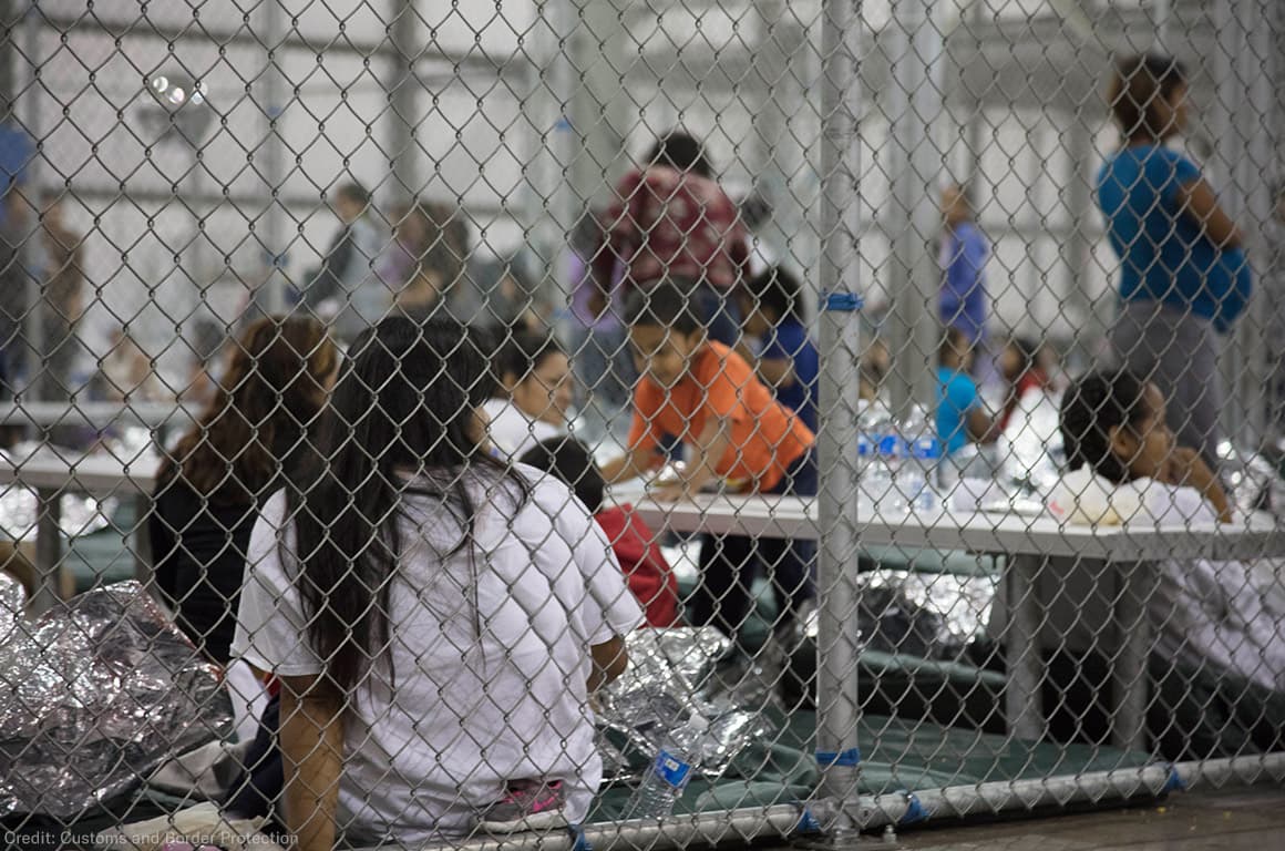 People — including children — sit behind chain-link fencing inside a U.S. immigration detention center.