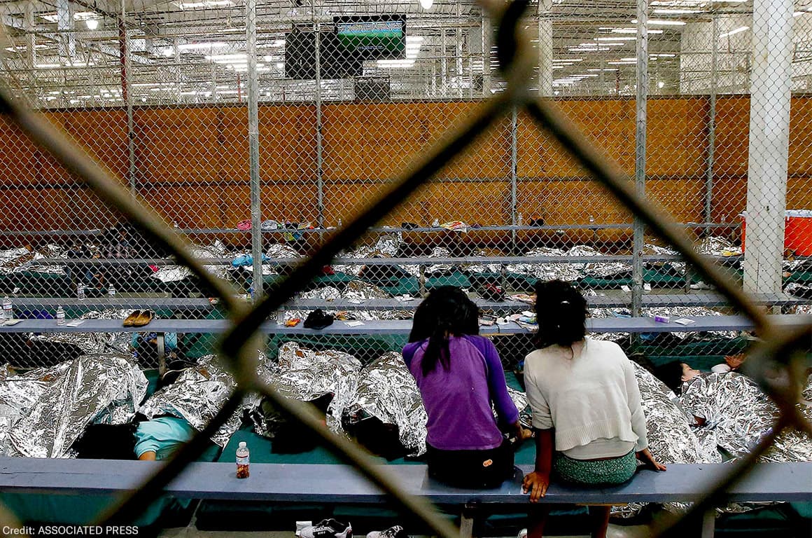 Two children sit together inside a fenced holding area filled with rows of mylar blankets.