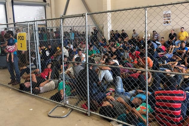 Rows of detained people behind fenced pens in a large indoor processing center.