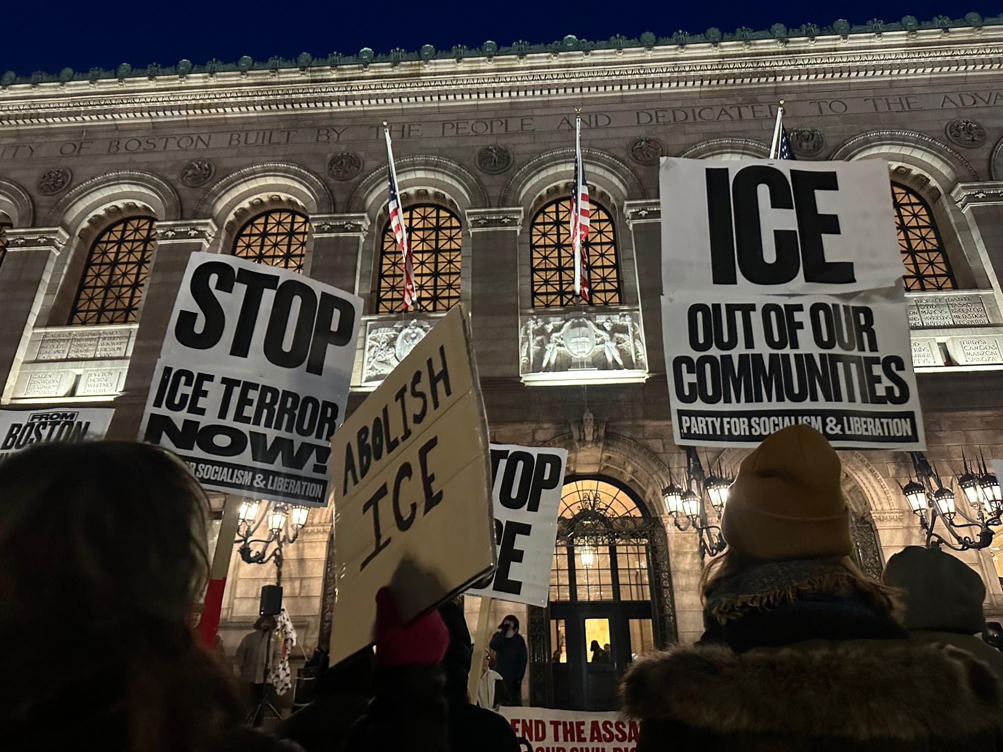 Evening rally outside a public library with 'Stop ICE terror now' and 'ICE out of our communities' signs.
