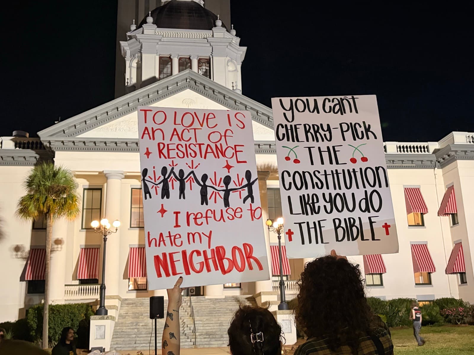 Night protest at the Florida State Capitol with signs reading 'To love is an act of resistance — I refuse to hate my neighbor' and 'You can't cherry-pick the Constitution like you do the Bible.'